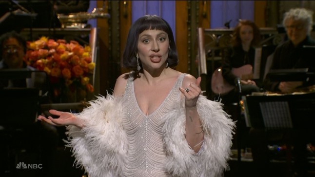 Lady Gaga in a glittery dress and white feather jacket on Saturday Night Live. She is speaking passionately to camera.
